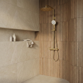 Nowoczesny prysznic ze złotym zestawem natynkowym Costa, w łazience z beżowymi płytkami i półką z kosmetykami.