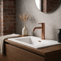 Nowoczesna łazienka z umywalką na drewnianej szafce z miedzianą baterią COSTA Veldman, betonową ścianą, cegłą w tle, lustrem oraz dekoracyjnym wazonem z kłosami.