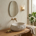 Naturalna łazienka z kamienną umywalką ALENA Veldman, drewnianym blatem, owalnym lustrem i złotą armaturą w ciepłym, przytulnym świetle.
