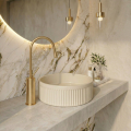 Elegancka łazienka z marmurową ścianą, okrągłym podświetlanym lustrem, złotą baterią i minimalistyczną ryflowaną umywalką beżową VERONA Veldman na blacie.