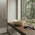 Minimalistyczna łazienka z owalną umywalką VERA w kolorze beżowy mat na drewnianym blacie, złotą baterią i widokiem na las przez okno.
